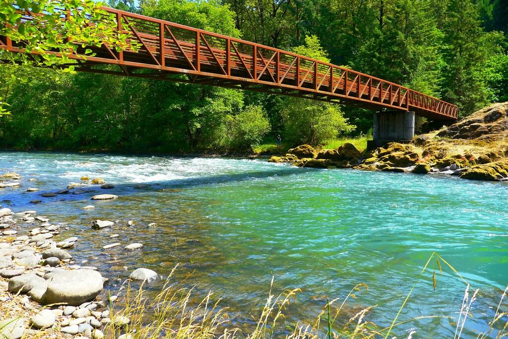 A rustic metal bridge spans a vibrant turquoise river in a sunny forest.