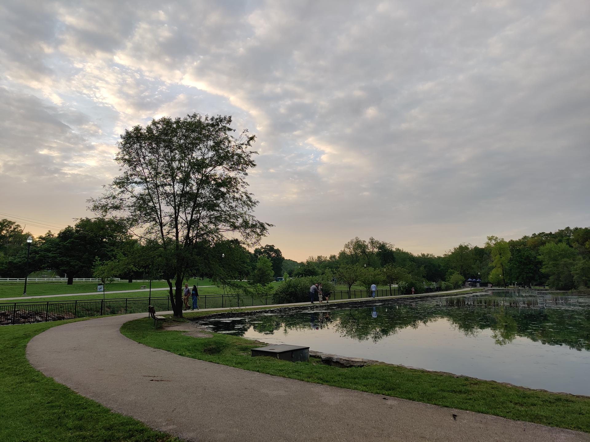 A paved path curves beside a calm lake under a cloudy sky at dusk.