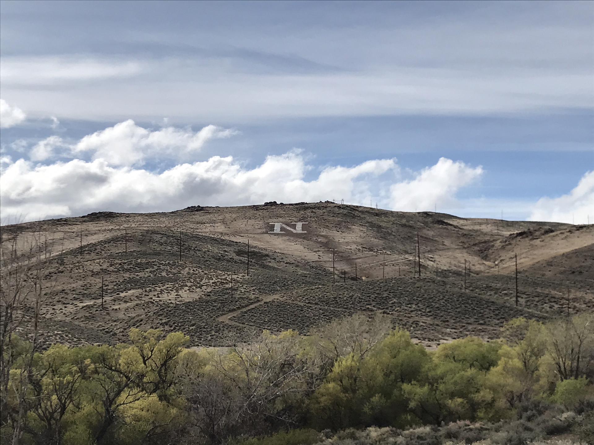 Large white letter "N" on a dry hillside with power lines and green trees.