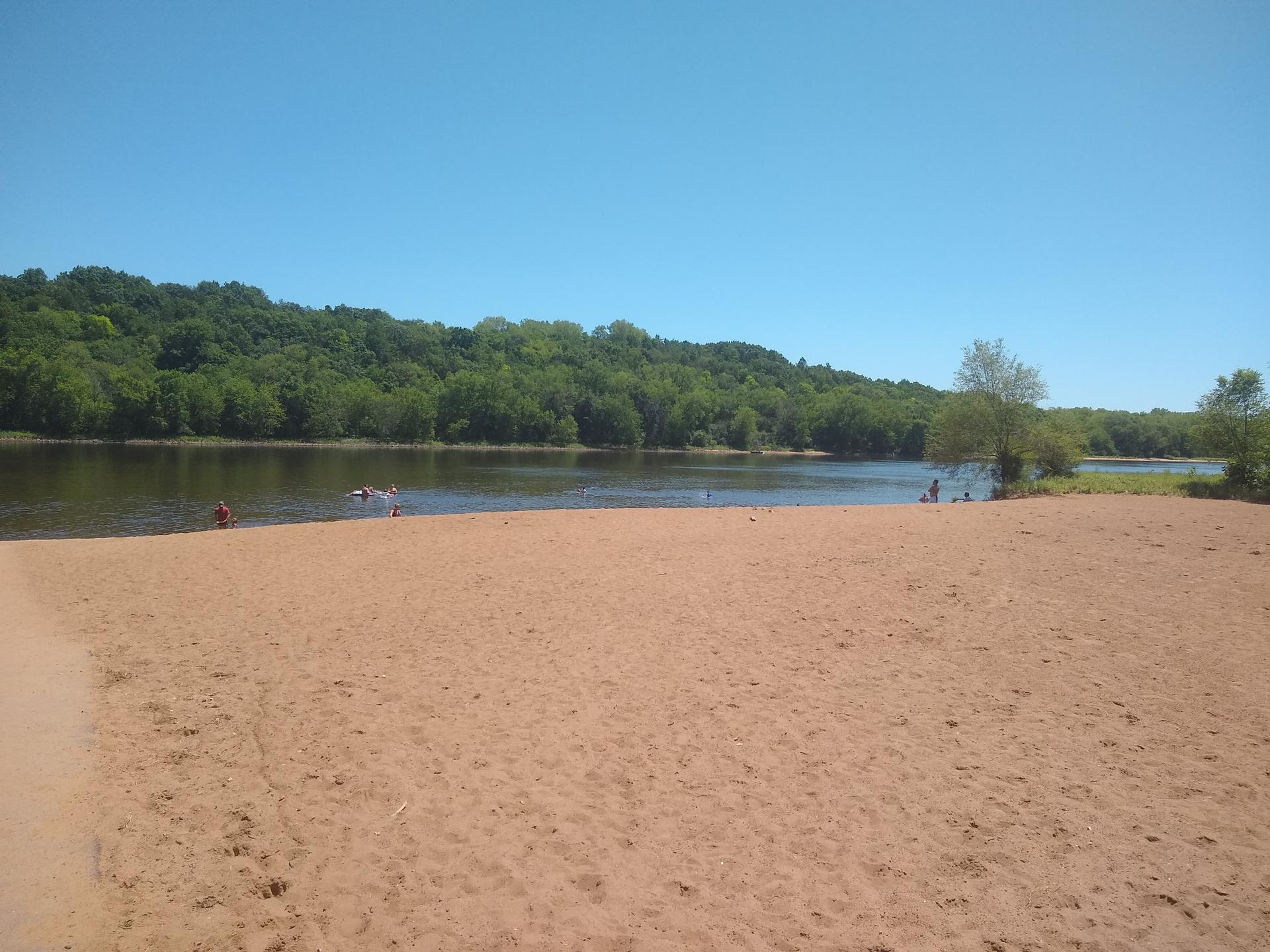 A wide sandy beach next to a river, with distant forested hills under blue sky.