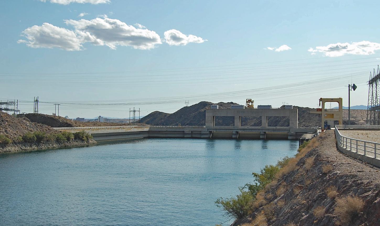 A wide canal flows towards a large concrete structure under a bright sky.