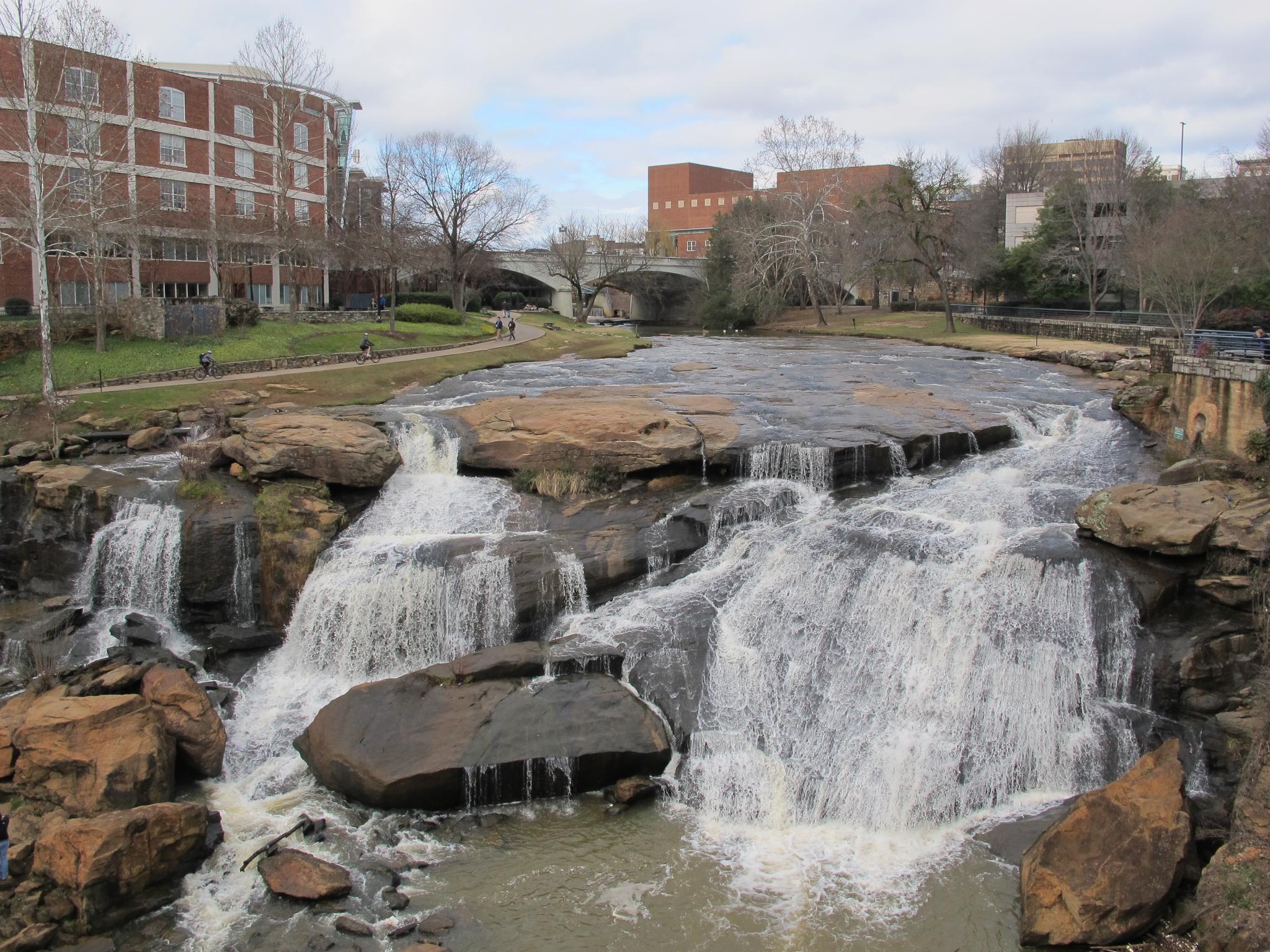 Wide waterfall over rocks, with buildings and a bridge in the background.
