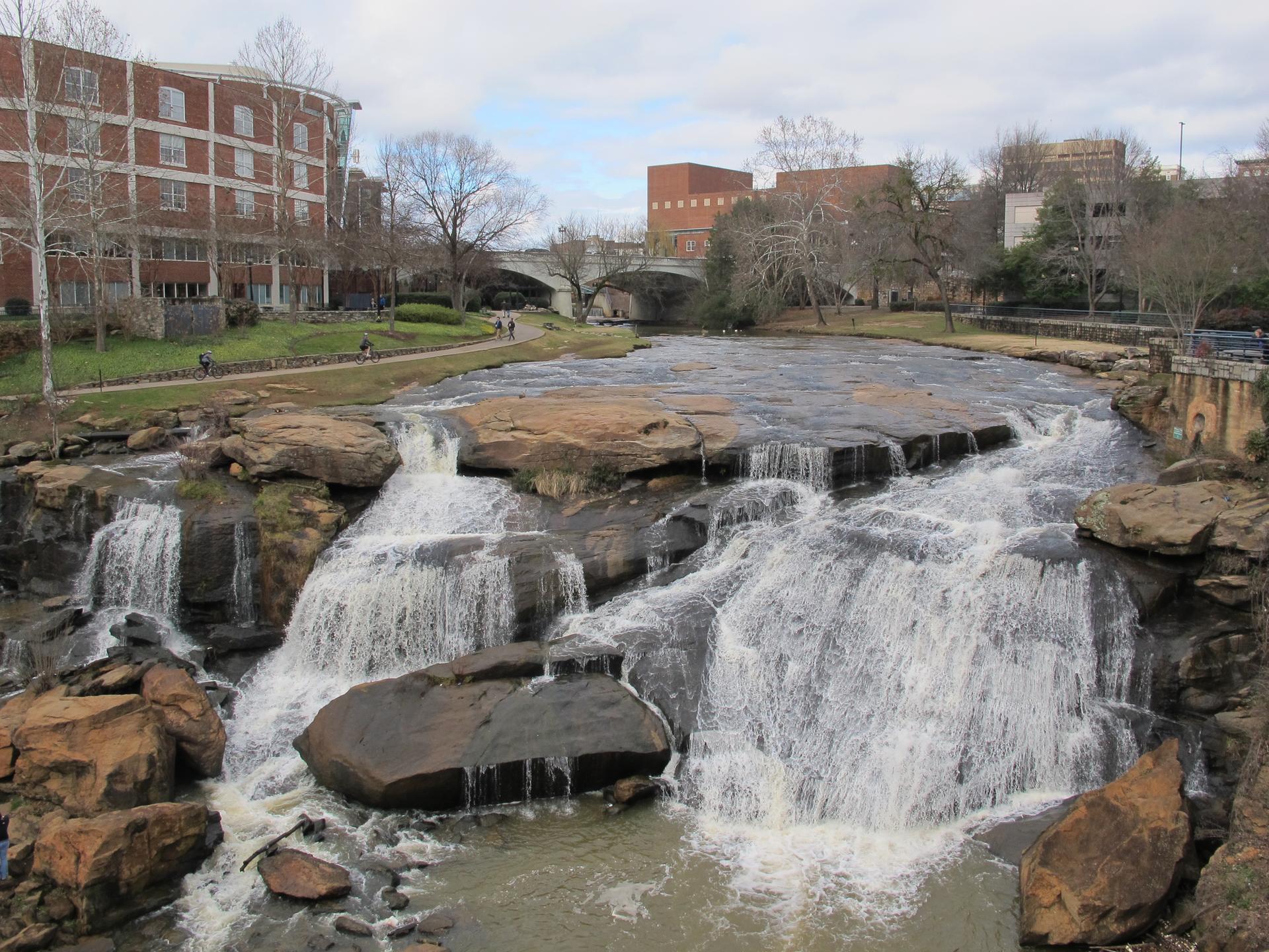 Wide waterfall over rocks, with buildings and a bridge in the background.