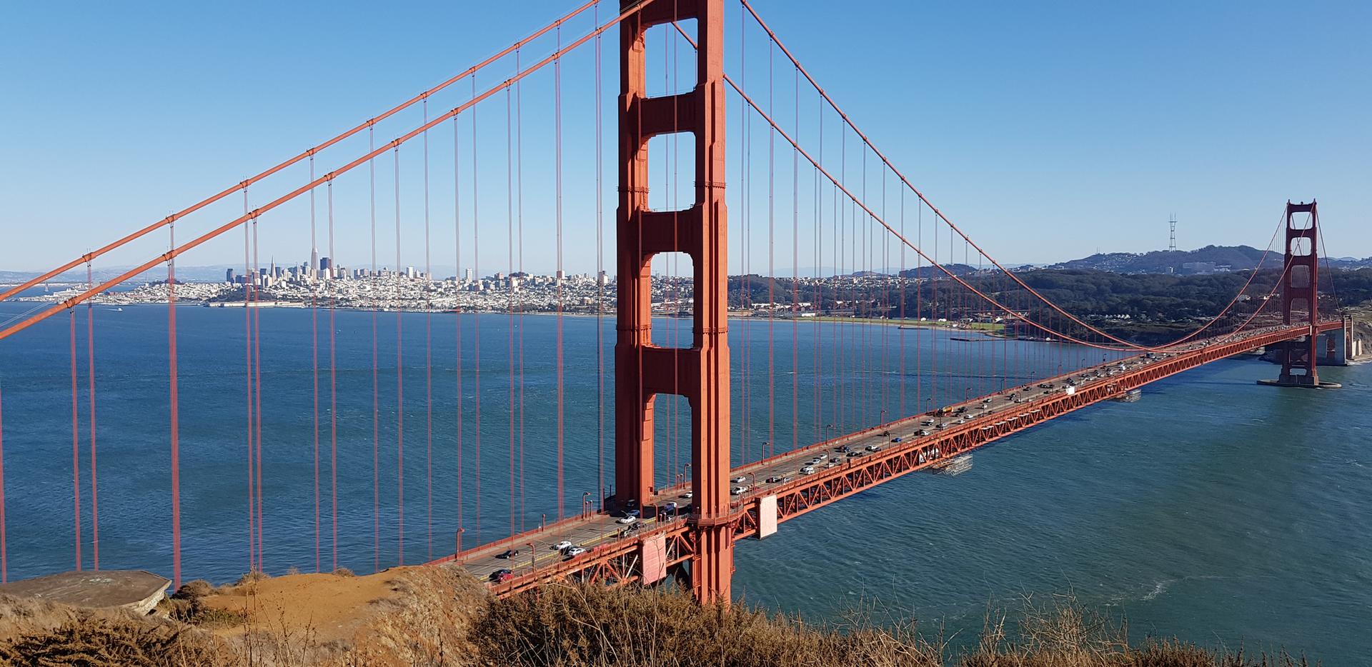 A large red suspension bridge spans blue water with a city skyline.