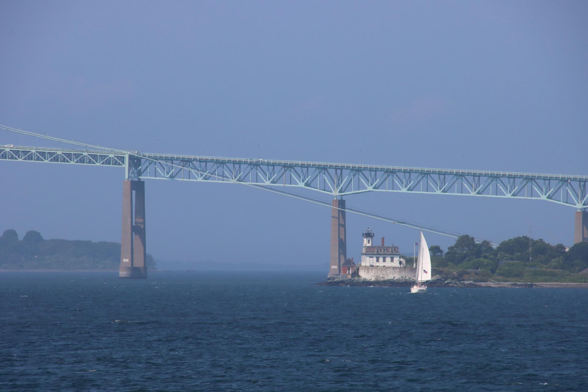 A large bridge spans over water with a sailboat passing a small island building.