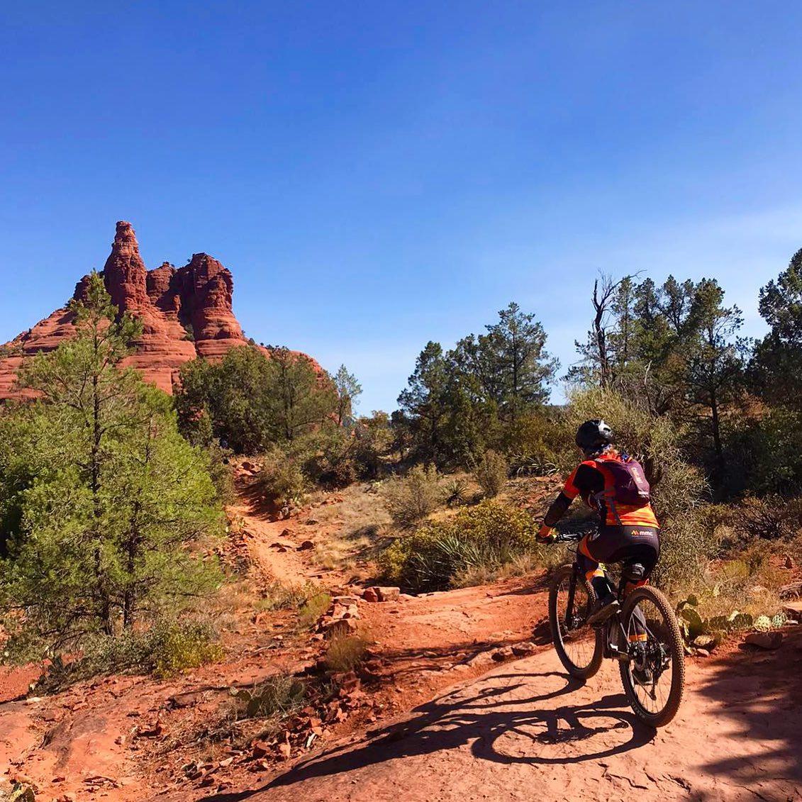 Mountain biker on a red dirt trail with large red rock formations.