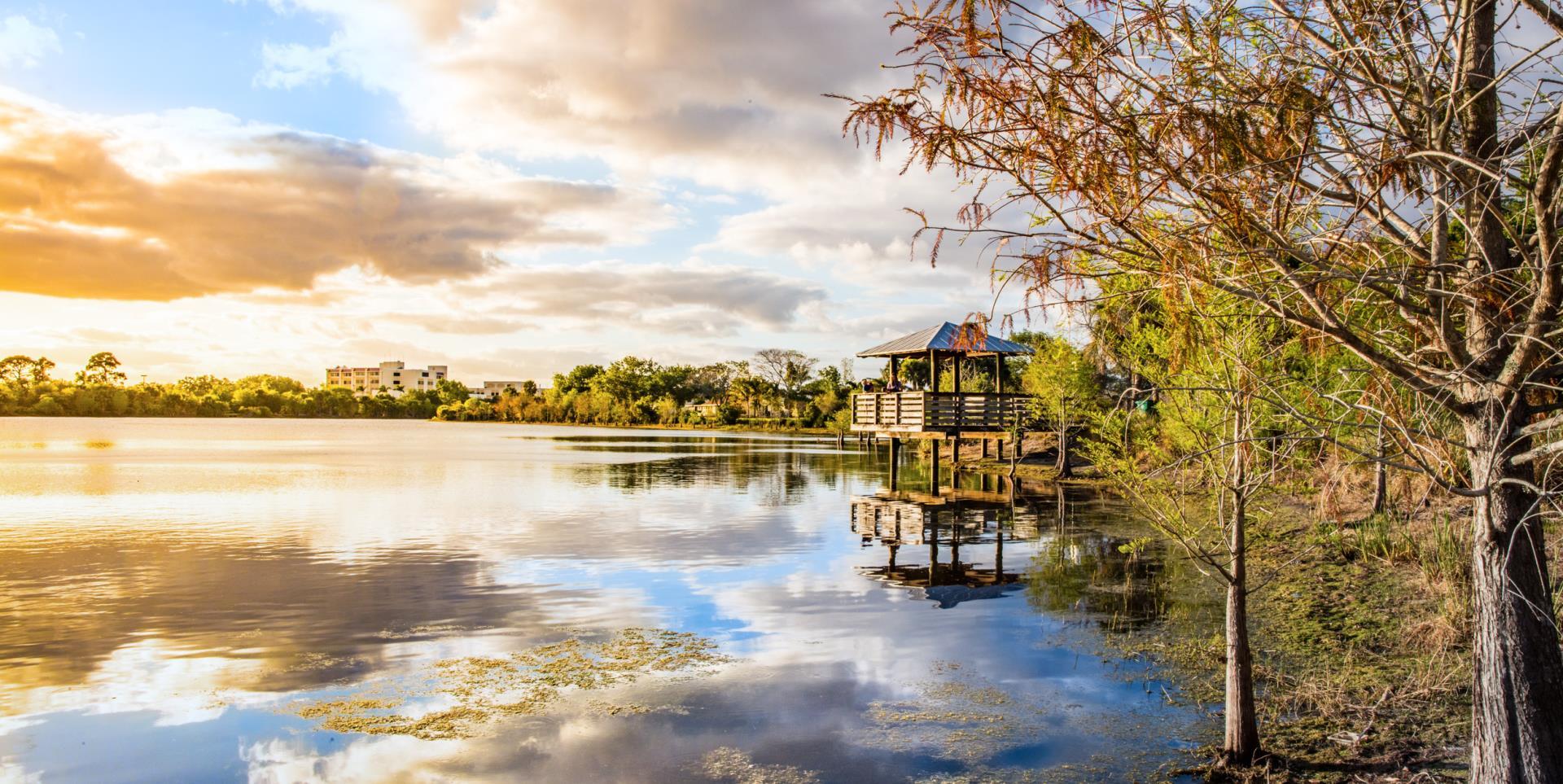 A wooden gazebo over a calm lake at golden hour, with reflections and trees.