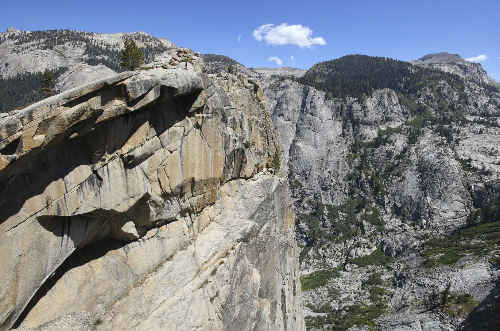 Dramatic rocky cliff face overlooking a vast mountain valley under a blue sky.