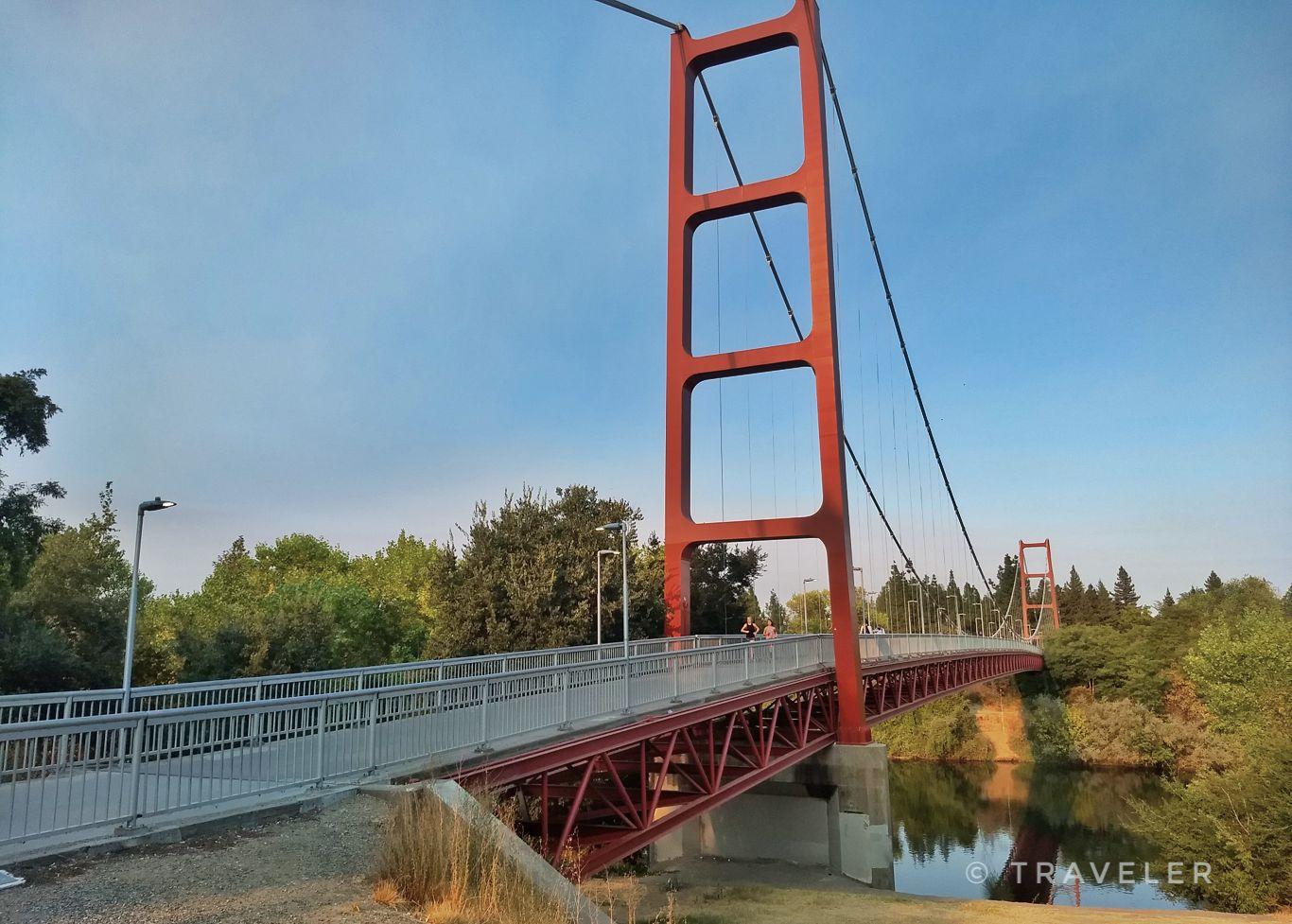 Red suspension bridge over calm water, surrounded by green trees, bright day.