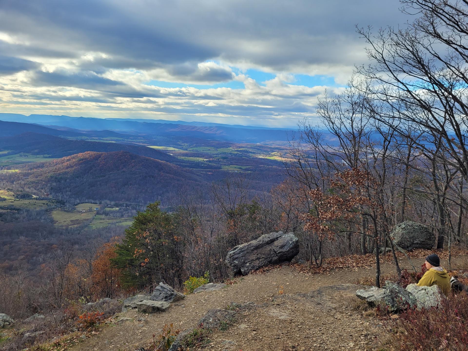Person sitting on a rocky overlook, viewing a vast mountain valley under cloudy skies.