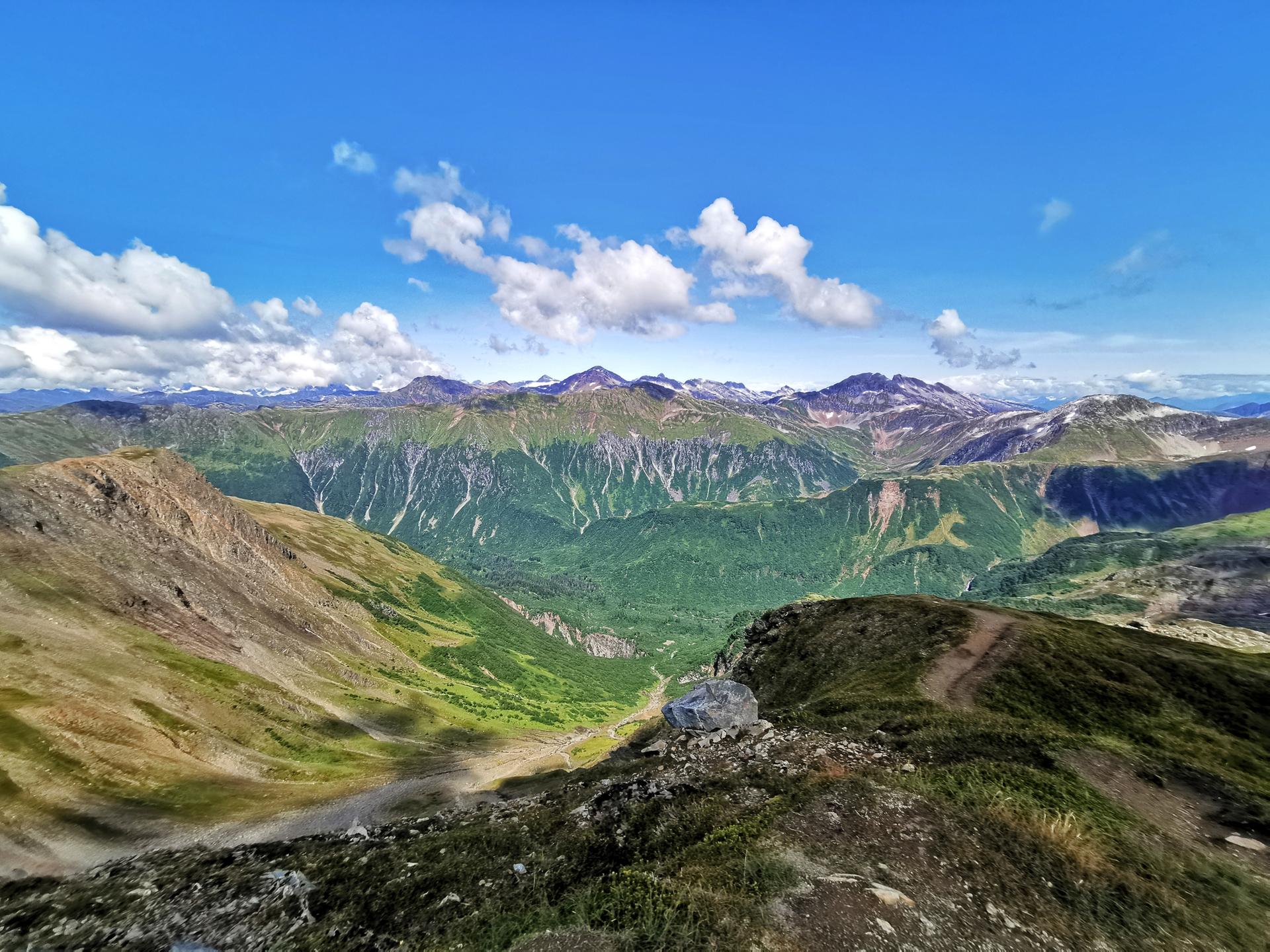 Expansive mountain range with green valleys under a bright blue sky with white clouds.