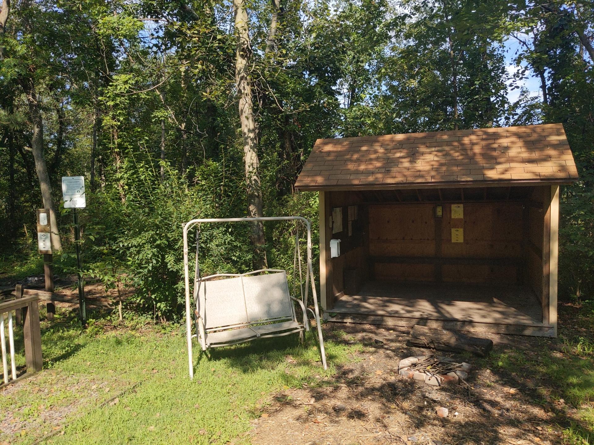 A rustic wooden shelter and a swing set in a sunny, wooded area.