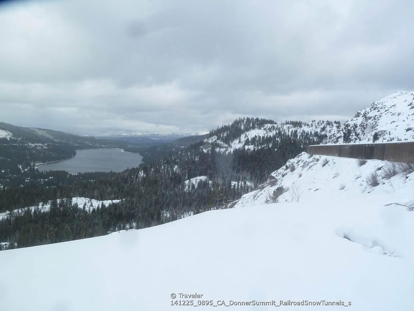 Vast snowy landscape with a distant lake, evergreen trees, and a snow shed.