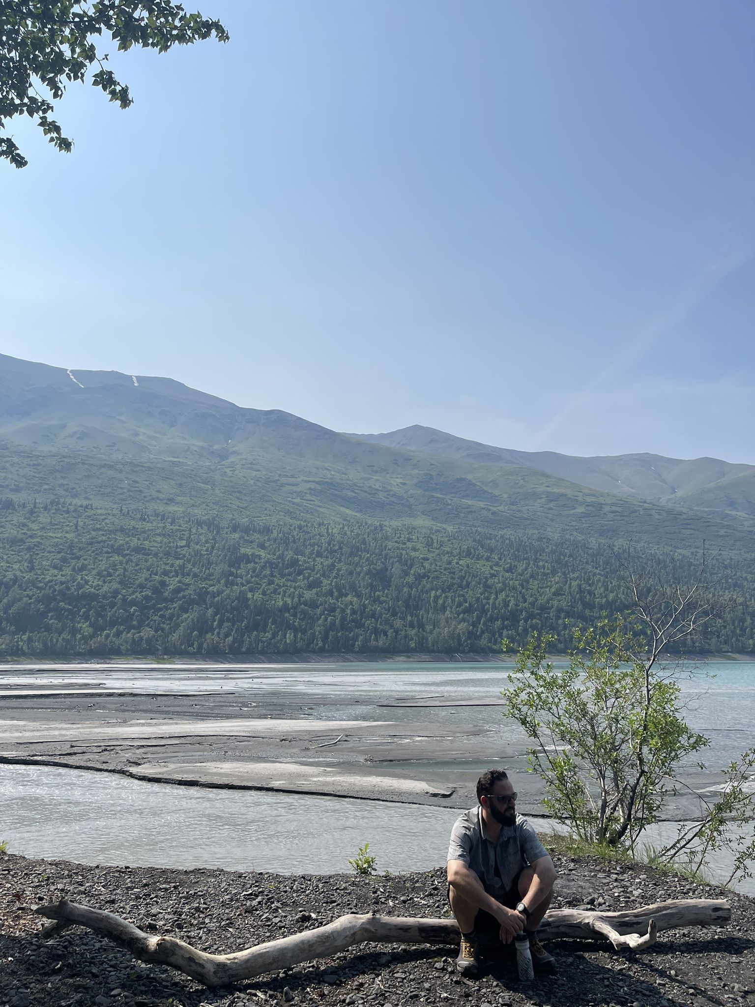A man sits on a dirt bank overlooking a wide river and forested mountains. - Eklutna Lakeside Trail