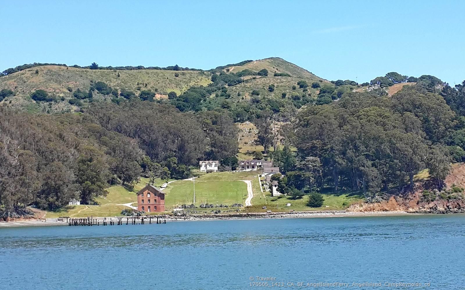 Waterfront buildings and a pier at the base of a green, tree-covered hill.