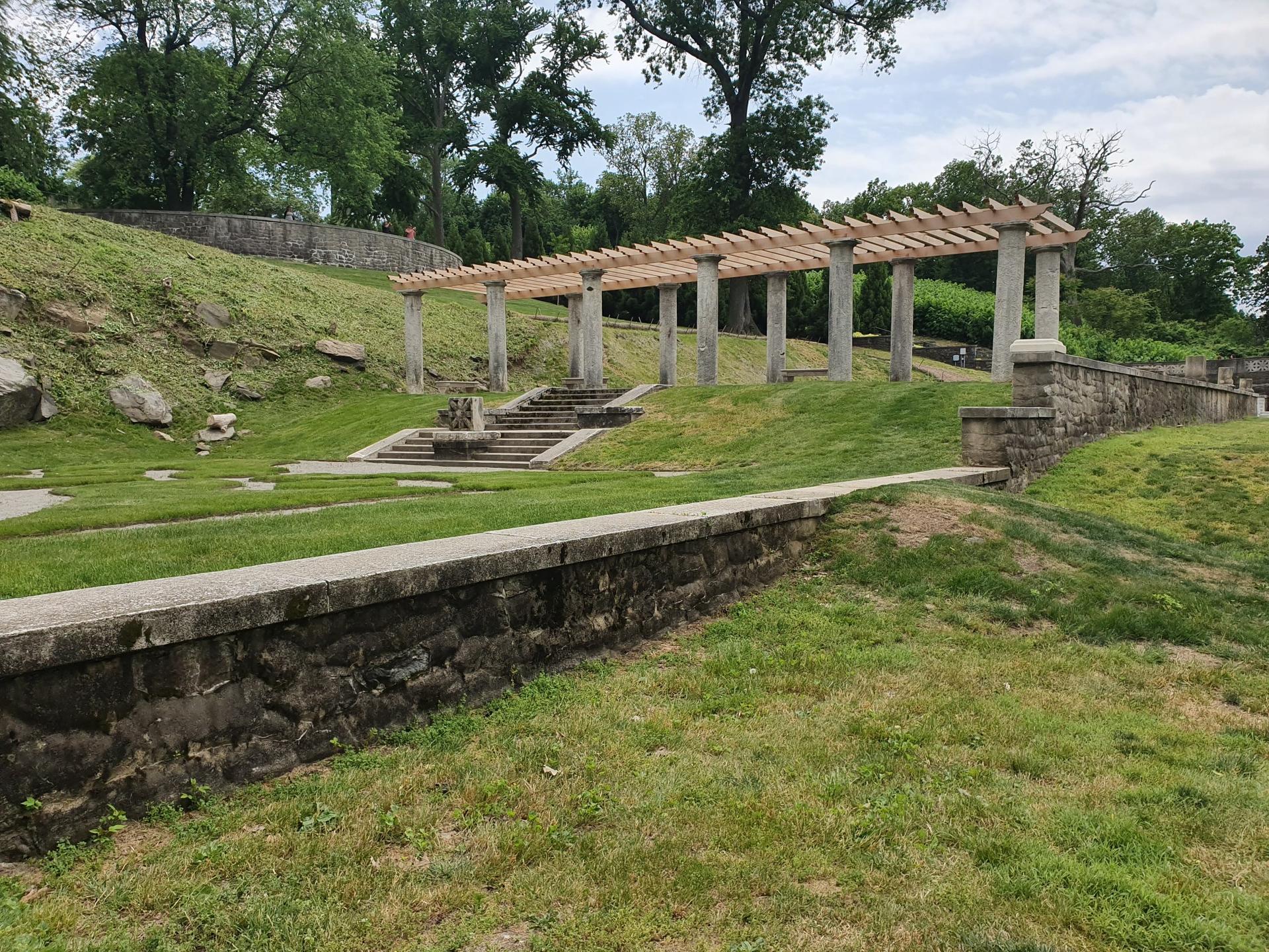Stone pergola on a grassy hill with steps, stone wall, and trees.