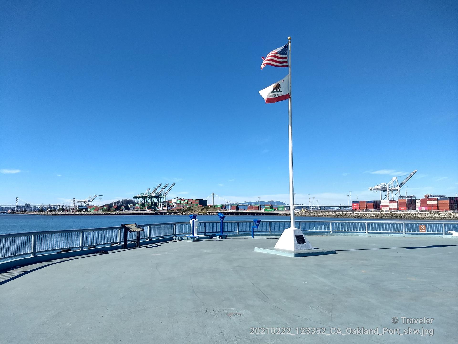 Two flags flying on a pier overlooking water and distant industrial cranes.