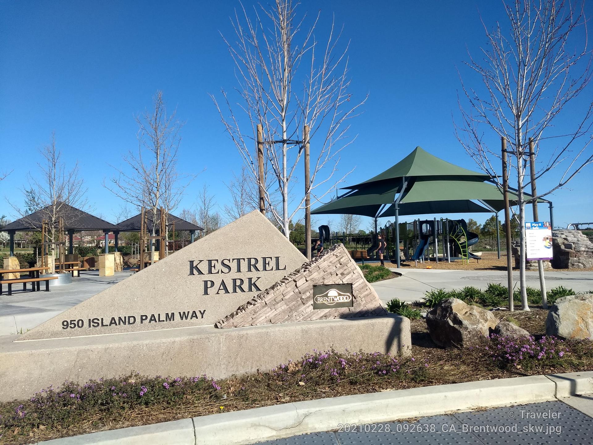 Park sign, bare trees, green canopies, and playground under bright sun.