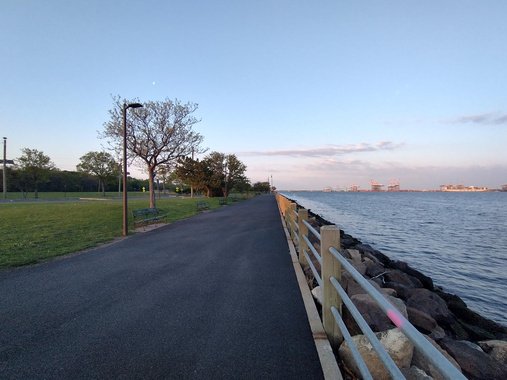 Paved path alongside calm water with trees under a soft sky.
