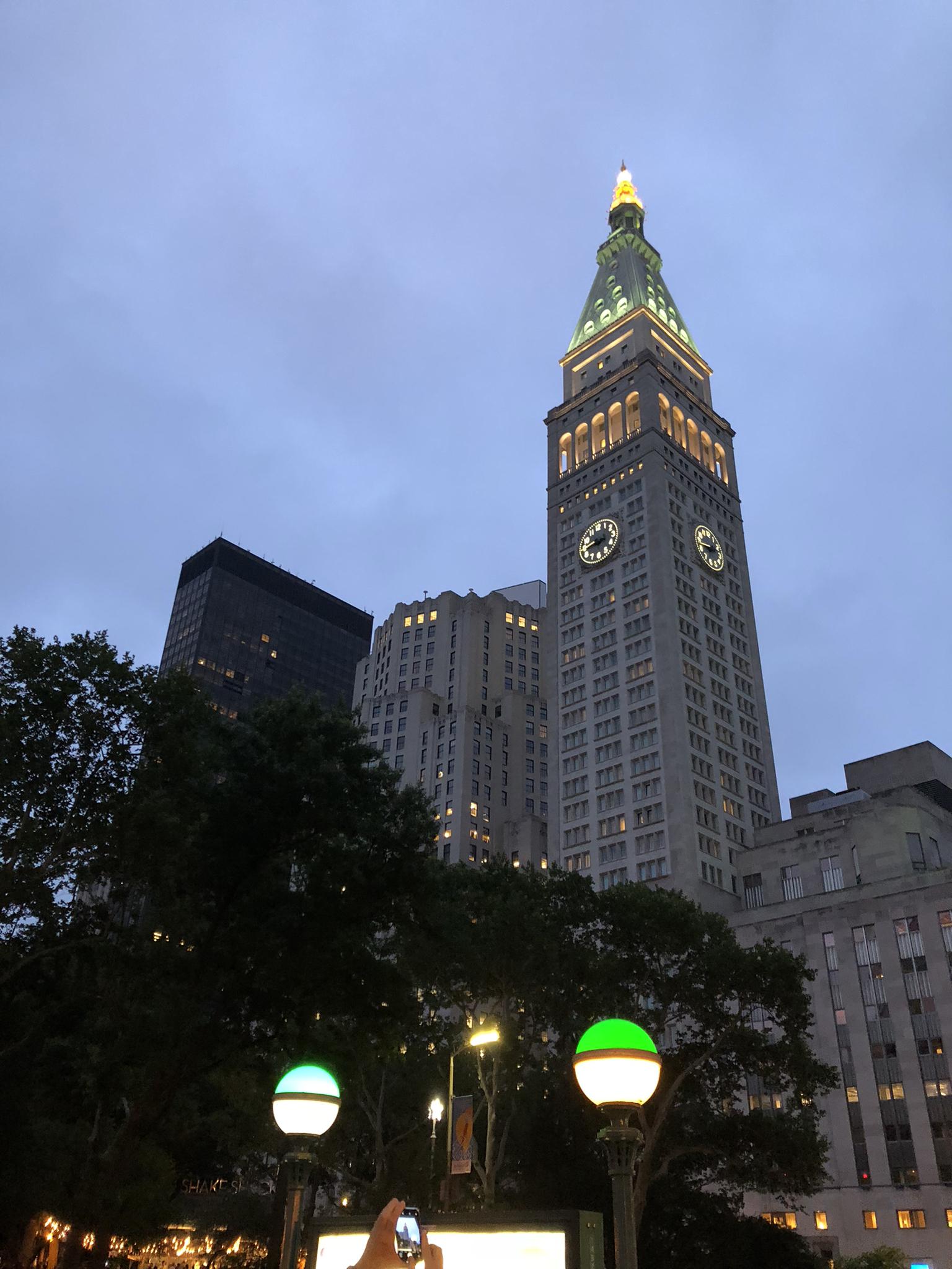 Illuminated clock tower building at dusk, with glowing green streetlights below.