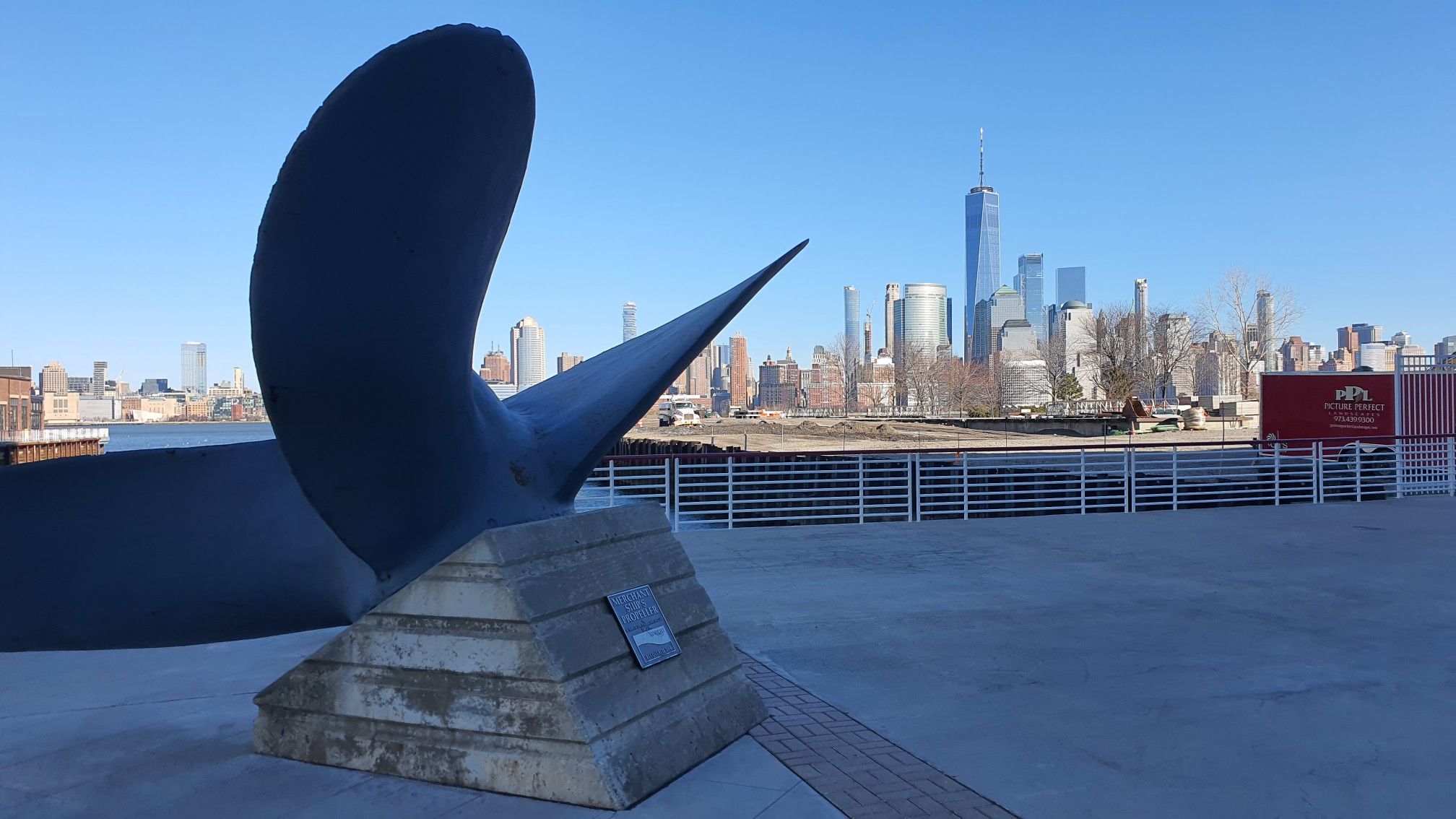 Large propeller sculpture on a paved area with a distant city skyline. - Hudson River Waterfront Walkway