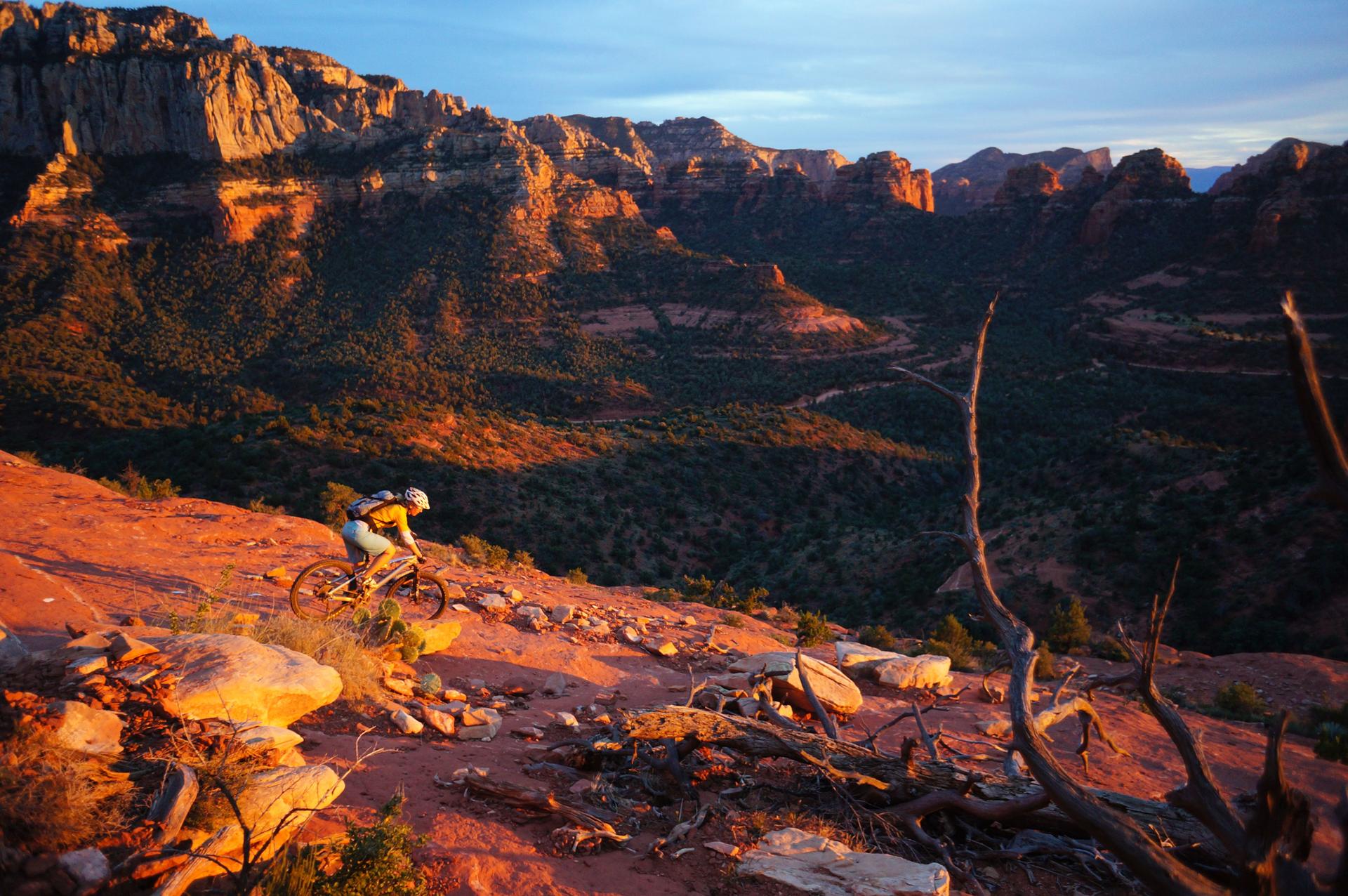 Mountain biker on a red trail at sunset, overlooking a vast, shadowed valley.