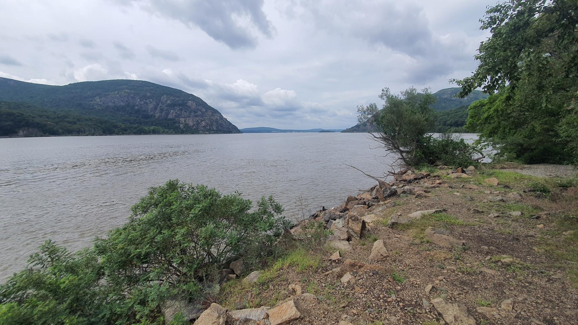 Wide river between mountains under cloudy sky, with rocky, vegetated bank.