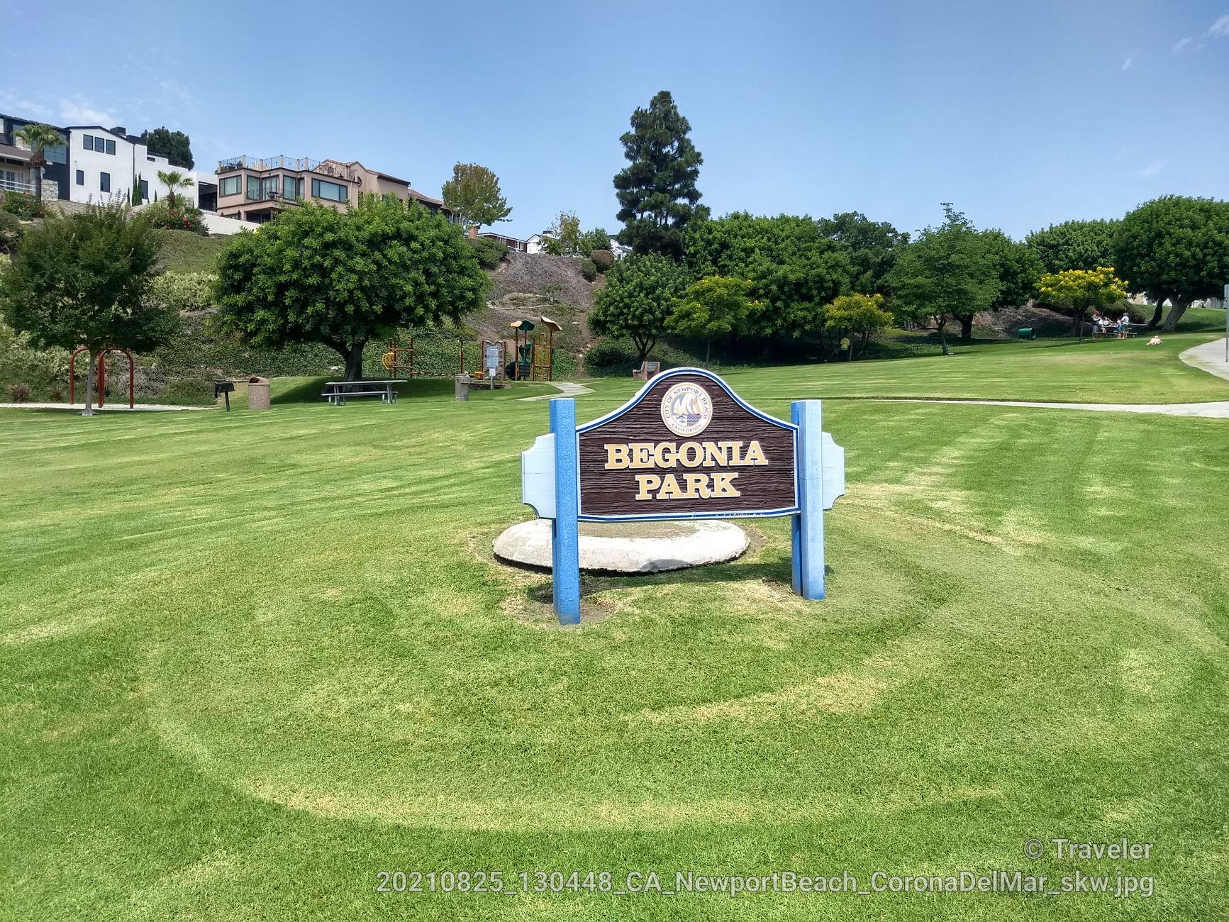 A park sign stands in a sunny, green grassy field with distant houses.