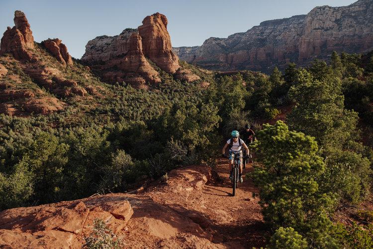 Two mountain bikers ride a narrow red trail through a dense green forest.