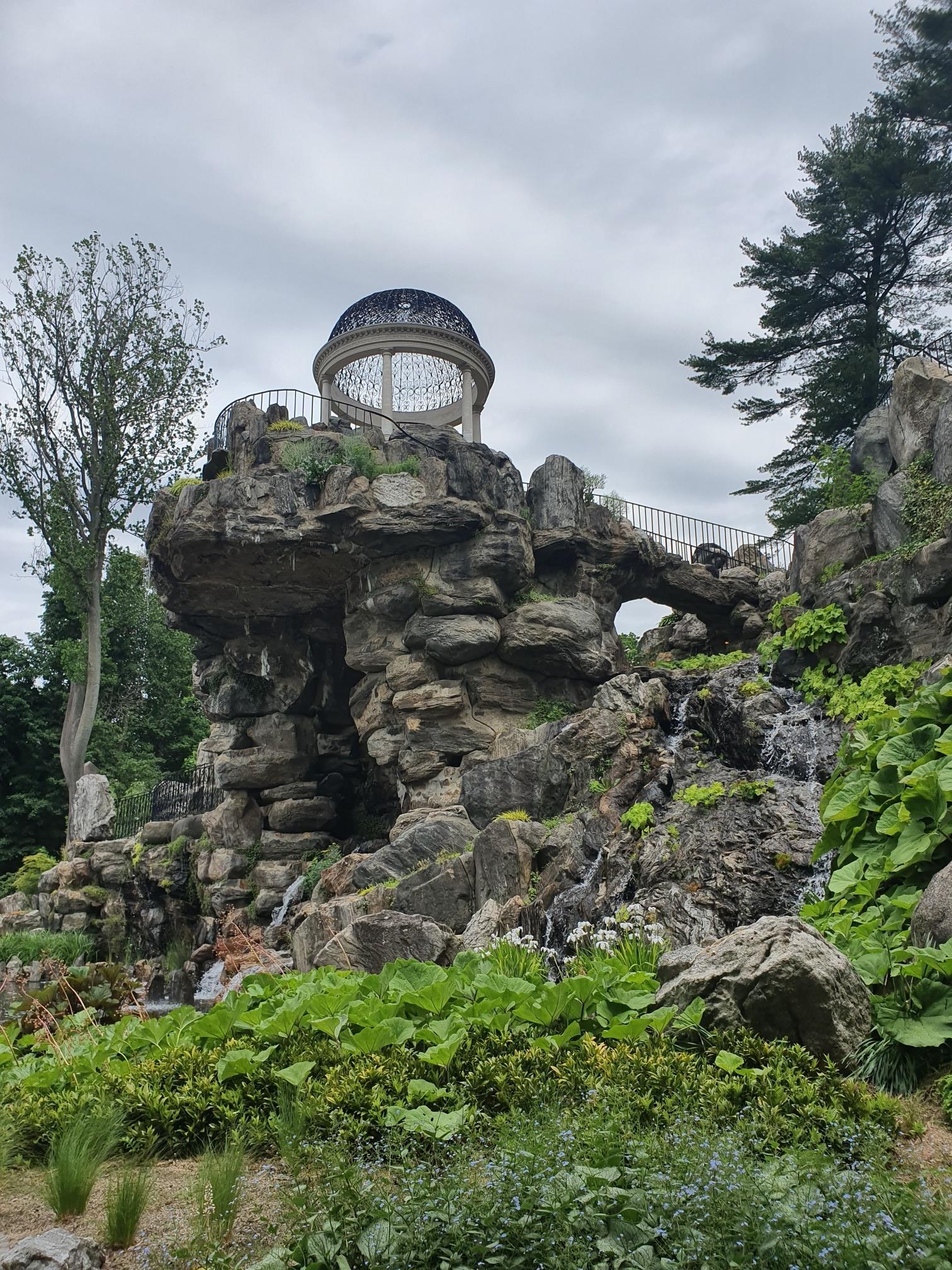 Stone grotto with waterfalls, ornate gazebo on top, lush plants, cloudy sky.