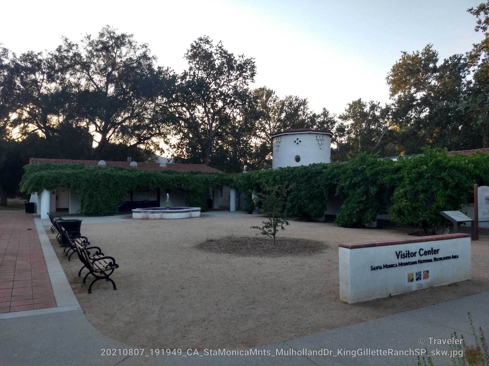 Building with a courtyard, fountain, and benches under trees in evening light.