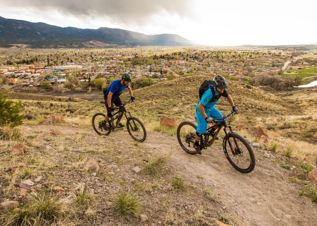 Two mountain bikers descend a dirt trail overlooking a valley town.