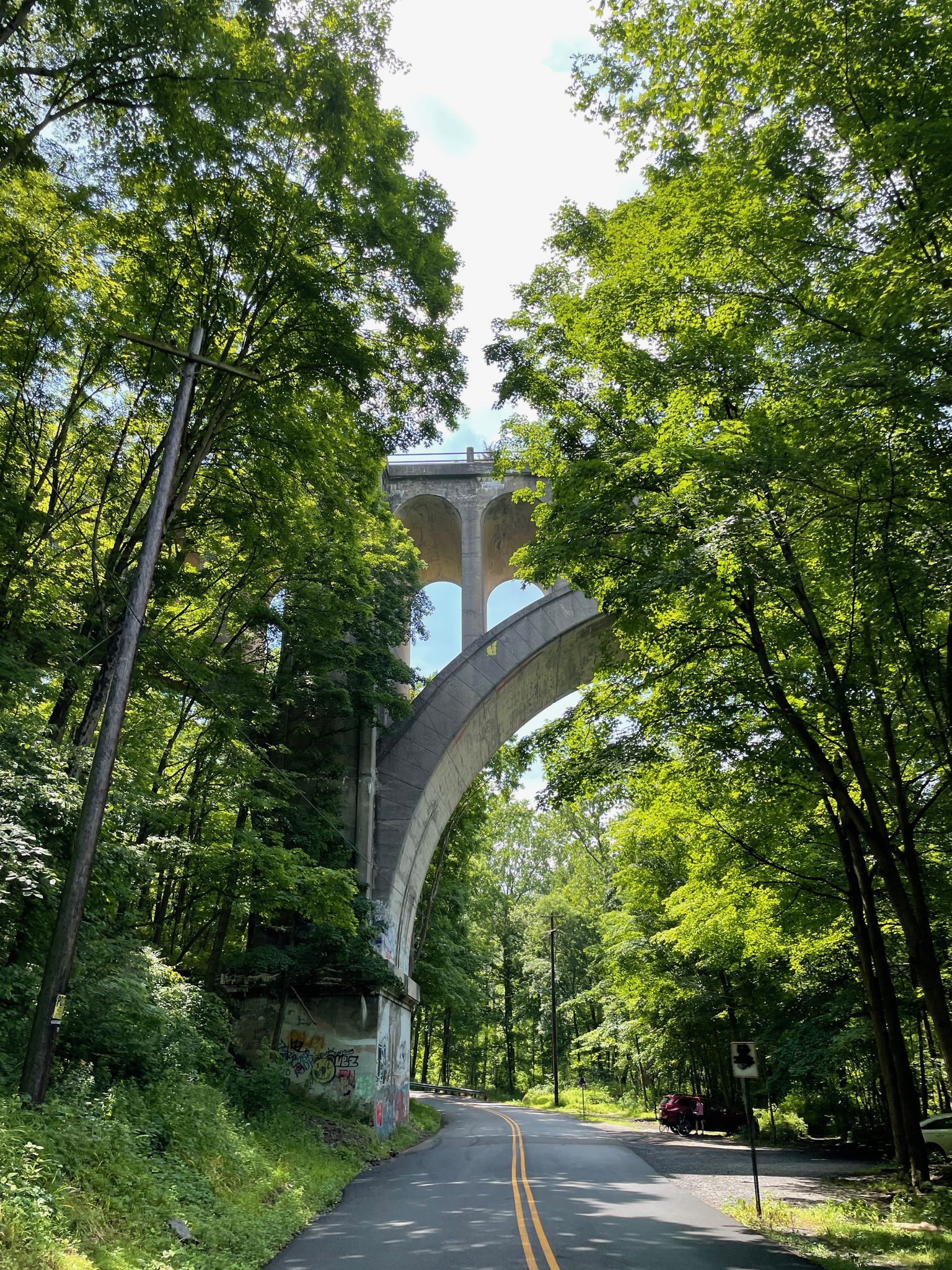 A large concrete arch bridge towers over a sunny road framed by green trees. - Paulinskill Viaduct – Paulinskill Valley Trail loop from Byram Township