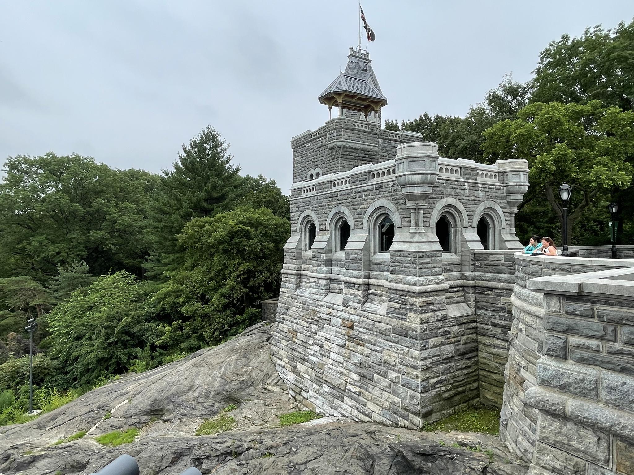 Stone castle-like structure on rocky hill, green trees, overcast sky.