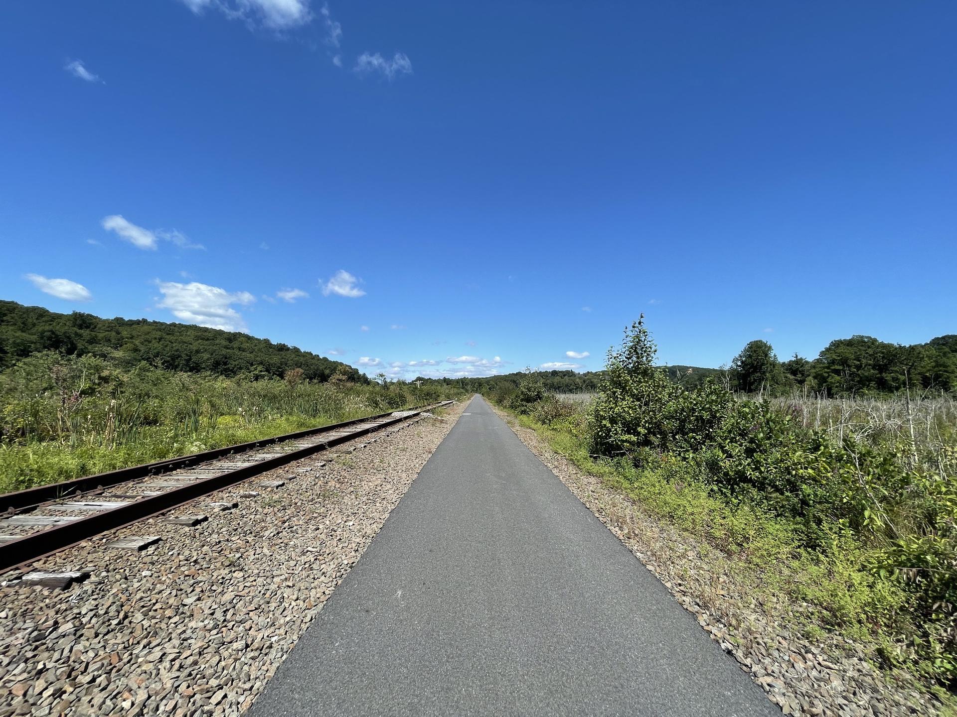 A paved path runs parallel to train tracks through green hills under blue sky.