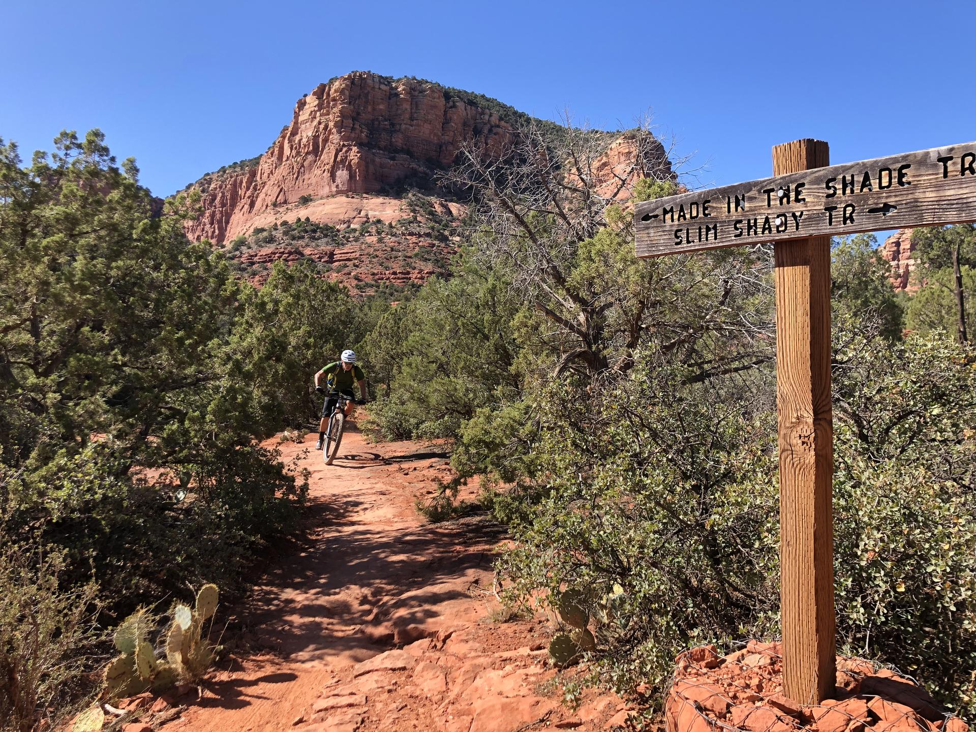 Mountain biker on a red trail next to a wooden sign, large mountain background.