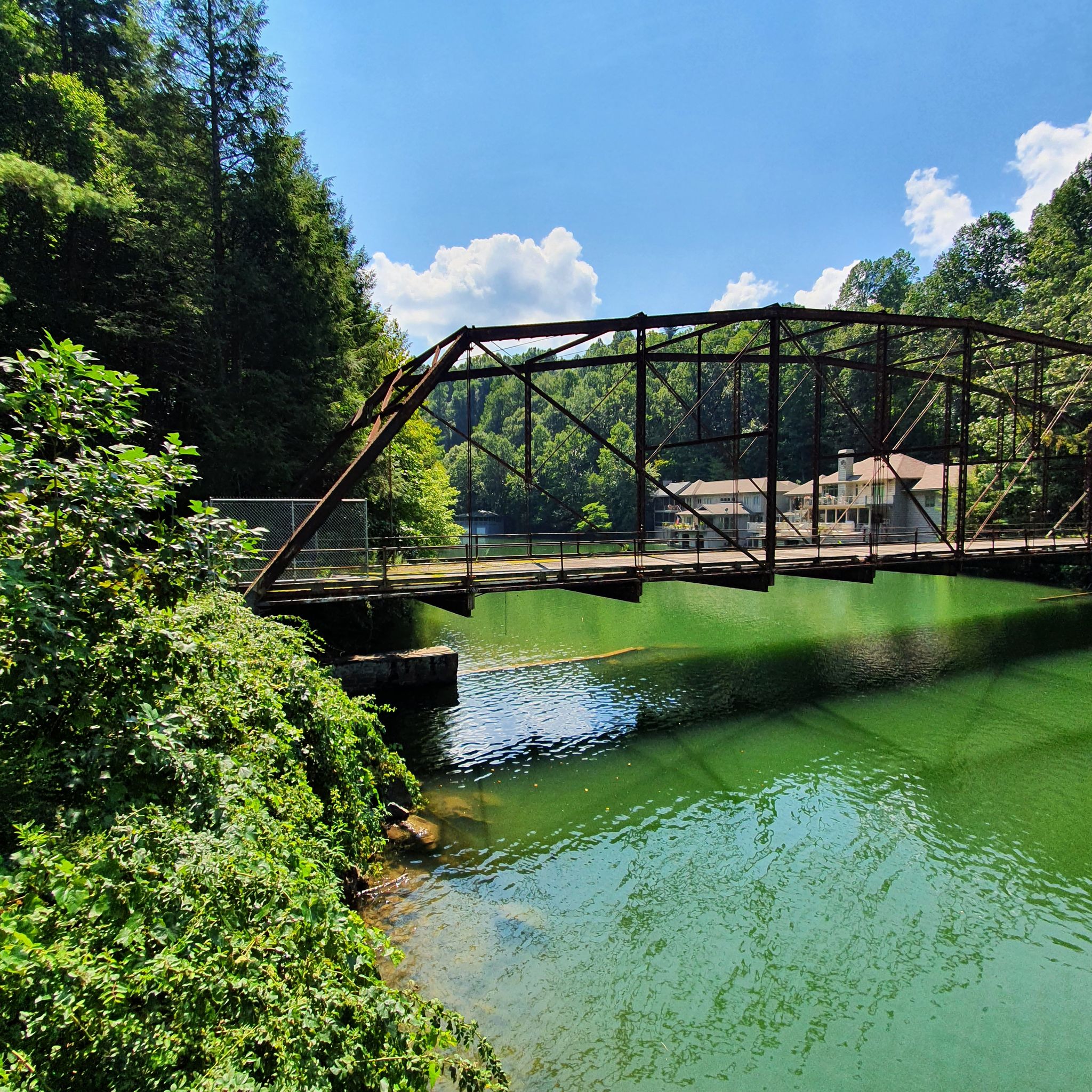 A metal truss bridge spans a vibrant green river under a bright sky. - Lake Summit Loop