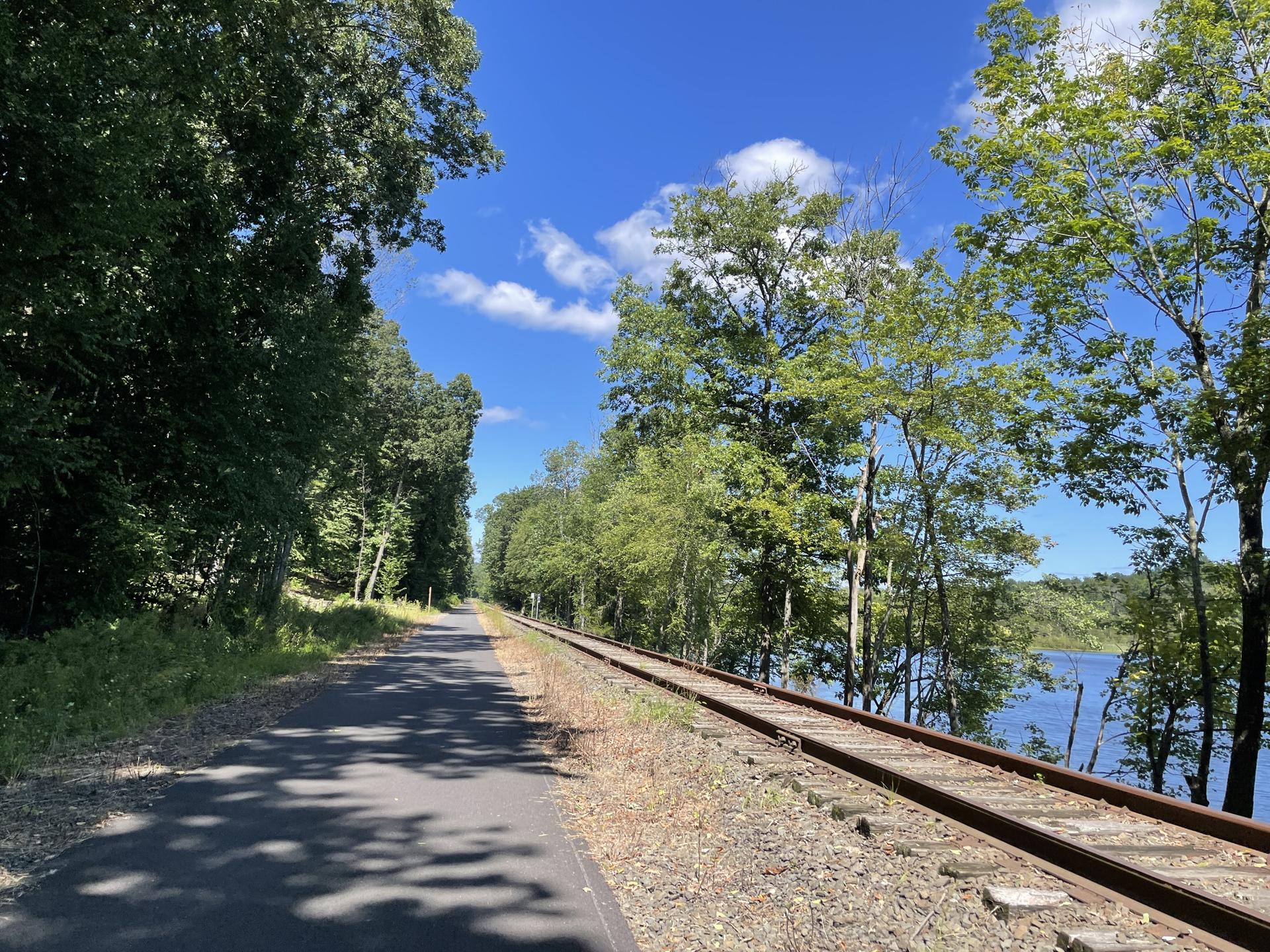 A paved path and train tracks run beside a lake, lined with green trees.