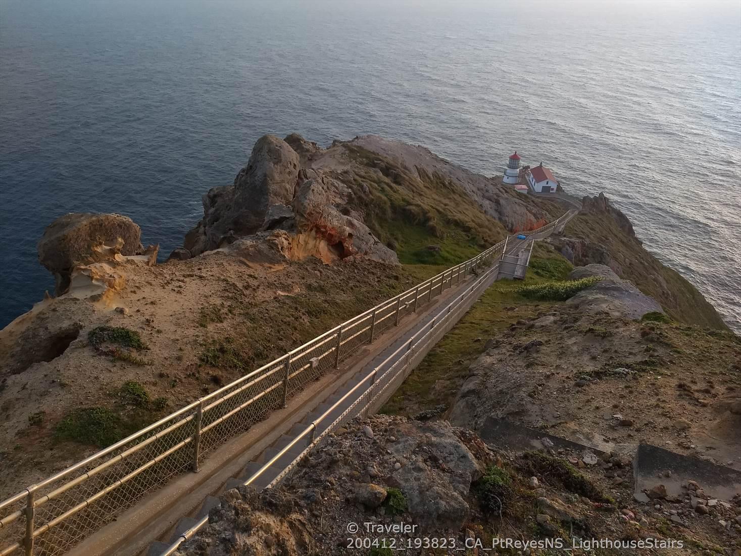 Fenced walkway leading to a lighthouse on a rocky cliff at sunset.