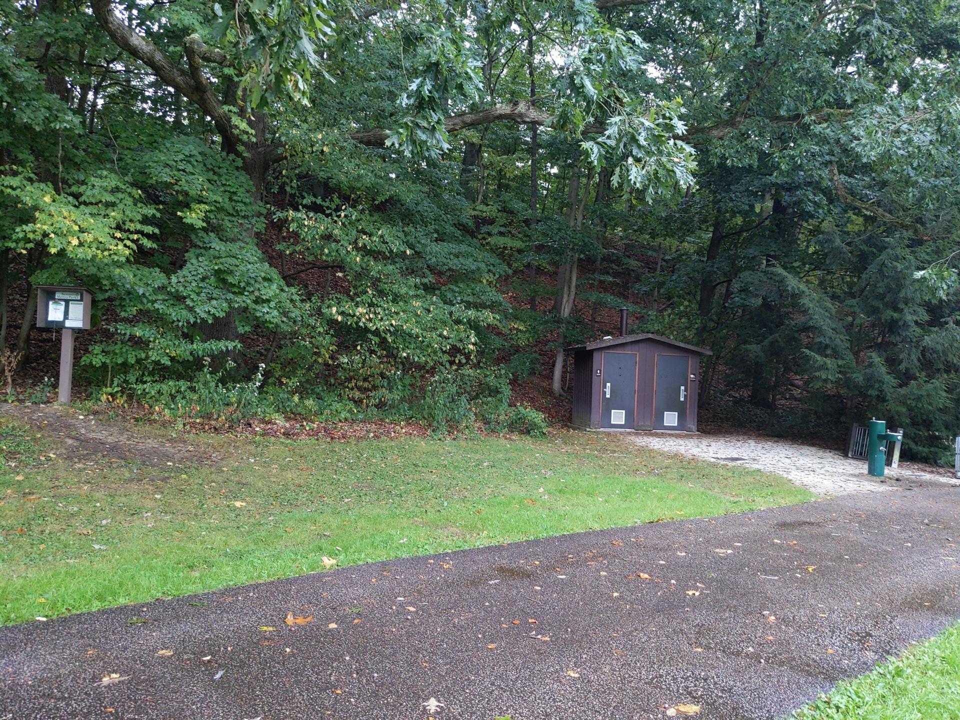 Small brown building at the edge of a paved path, surrounded by trees.