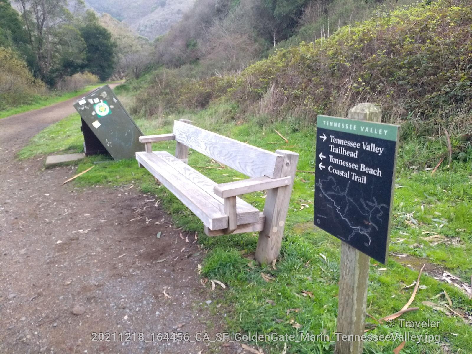 A wooden bench and trail sign on a dirt path, green hills beyond.