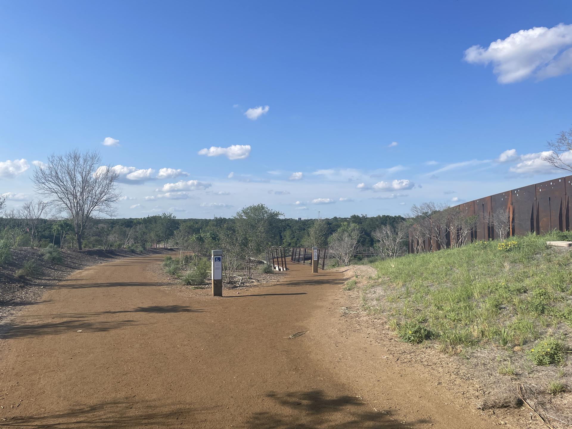 A wide dirt path under a bright blue sky, with trees and a tall fence.