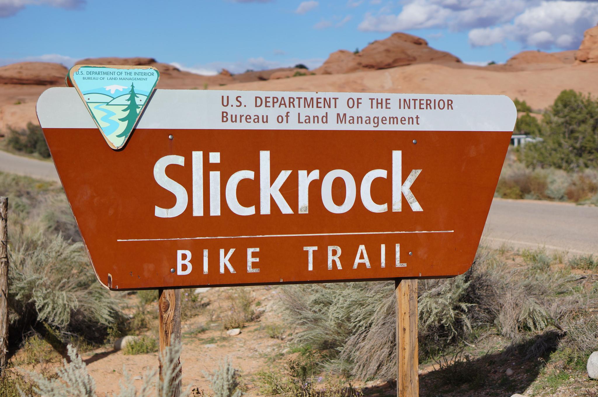 Brown sign for "Slickrock Bike Trail" with desert hills in background.