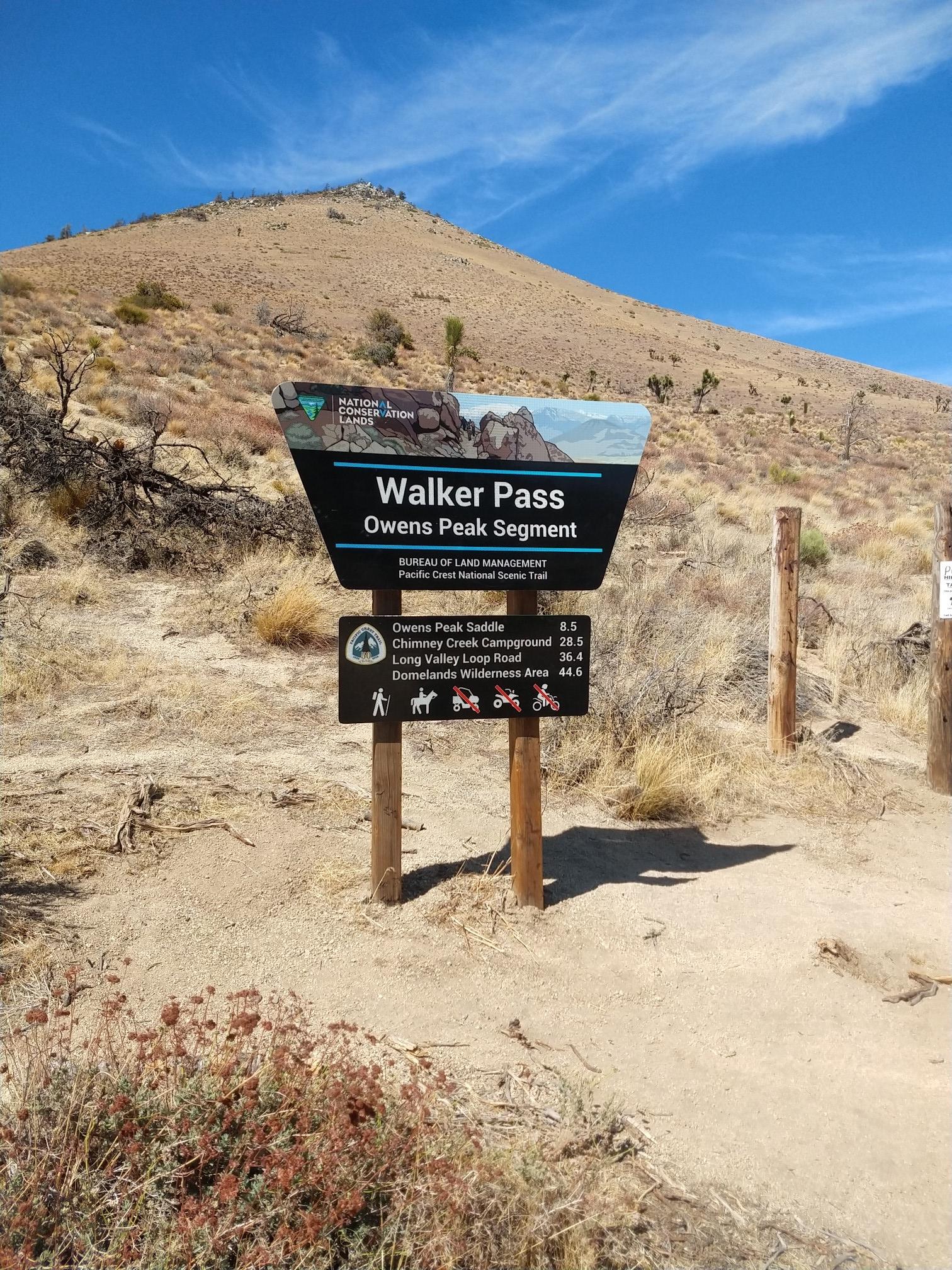 A trail sign stands on a dry, sunny hillside with a mountain behind.