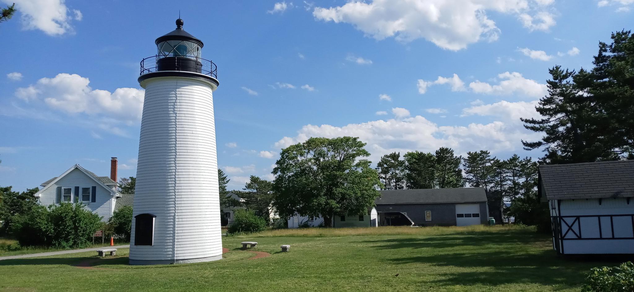 White lighthouse on a green lawn under a bright blue sky with scattered clouds.