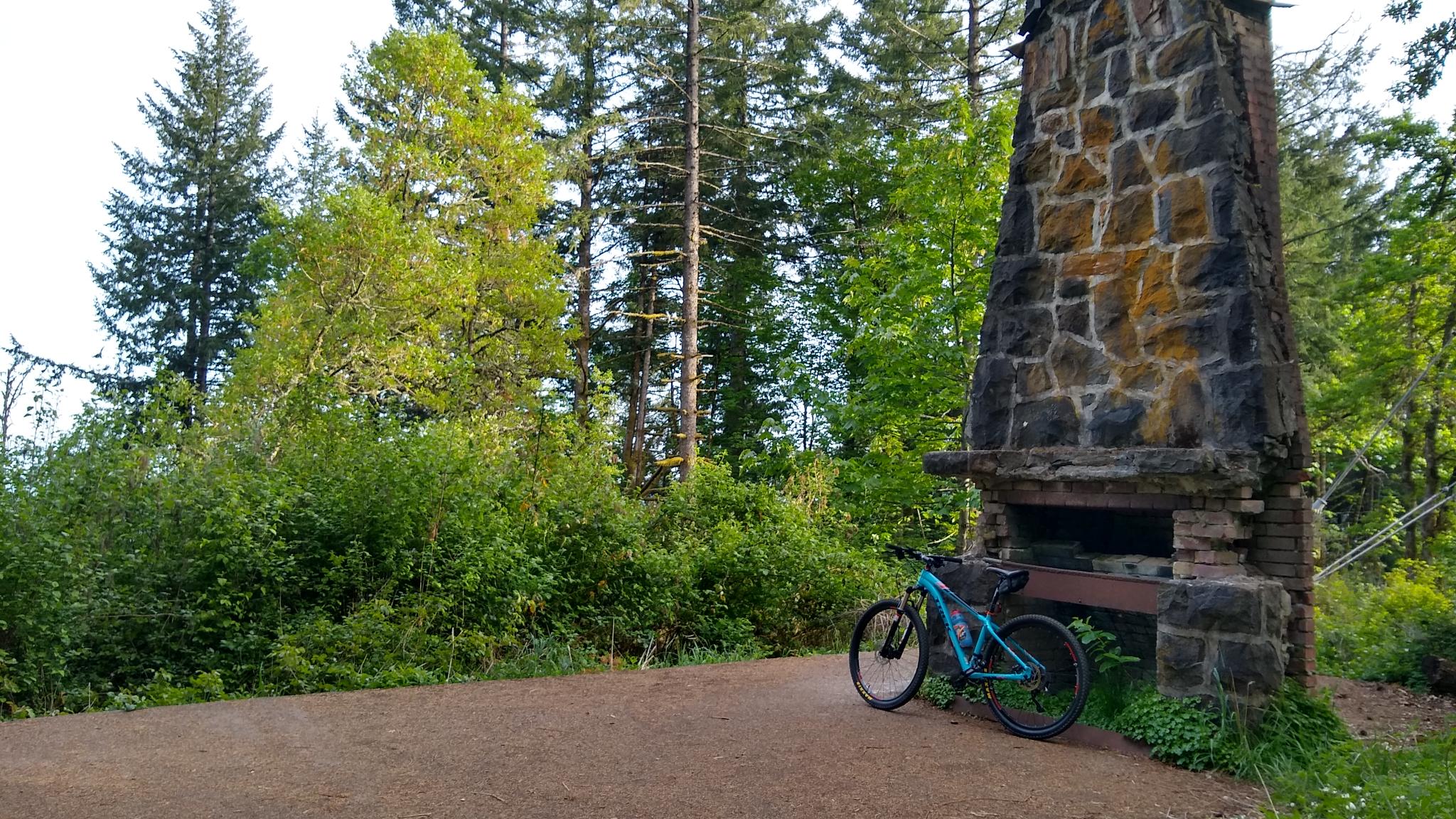 A blue mountain bike parked beside a stone chimney in a bright forest.