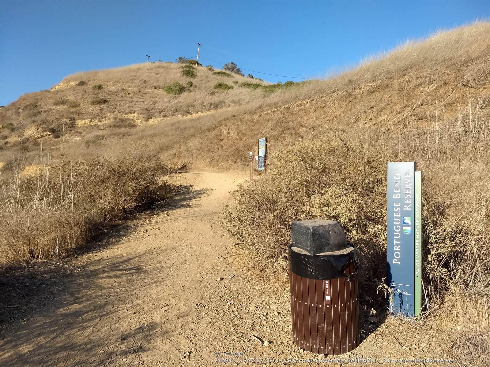 A dirt path winds up a dry, sunny hill with a trash can.
