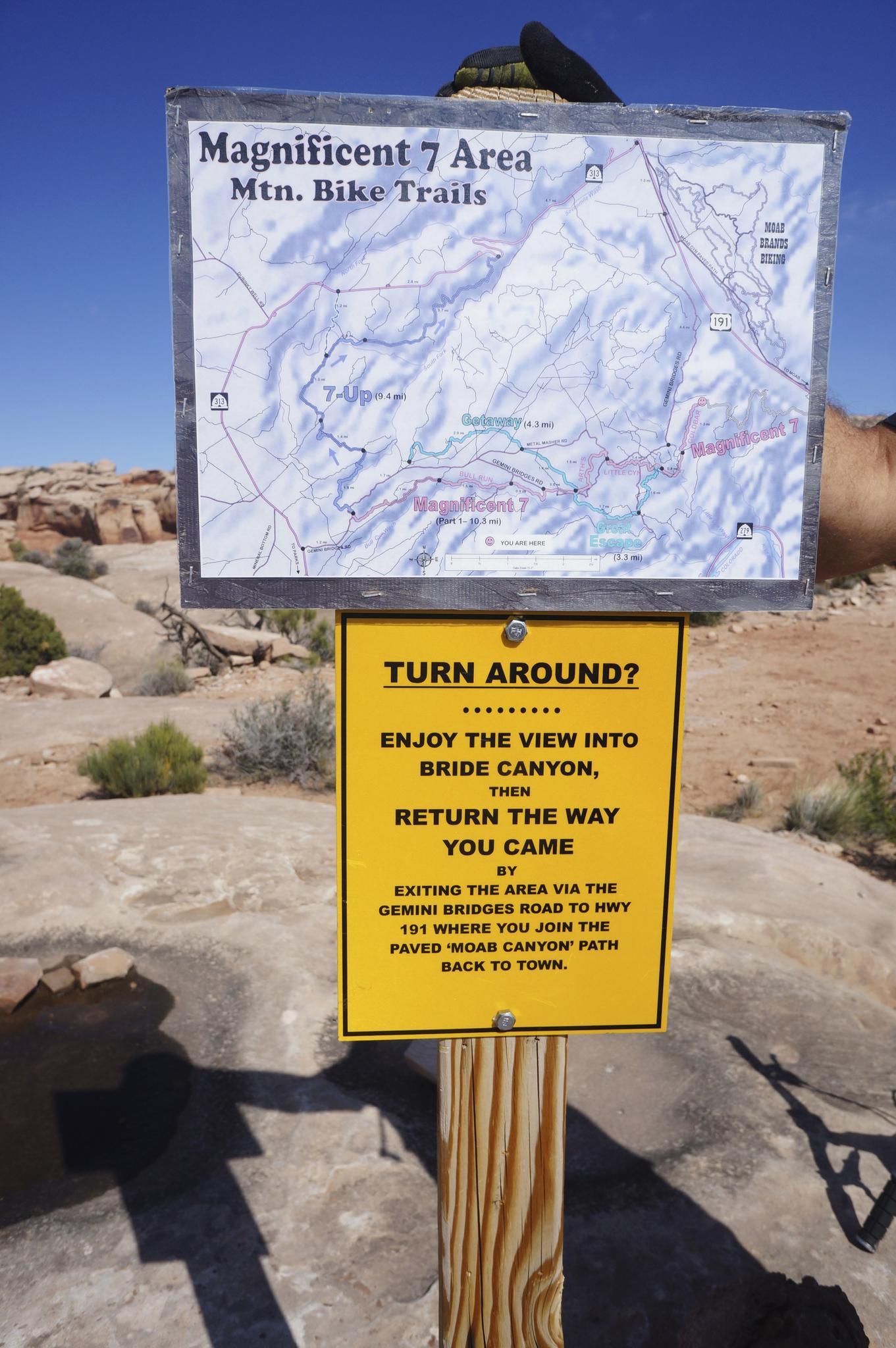 A trail map and warning sign on a wooden post in a rocky, arid environment.