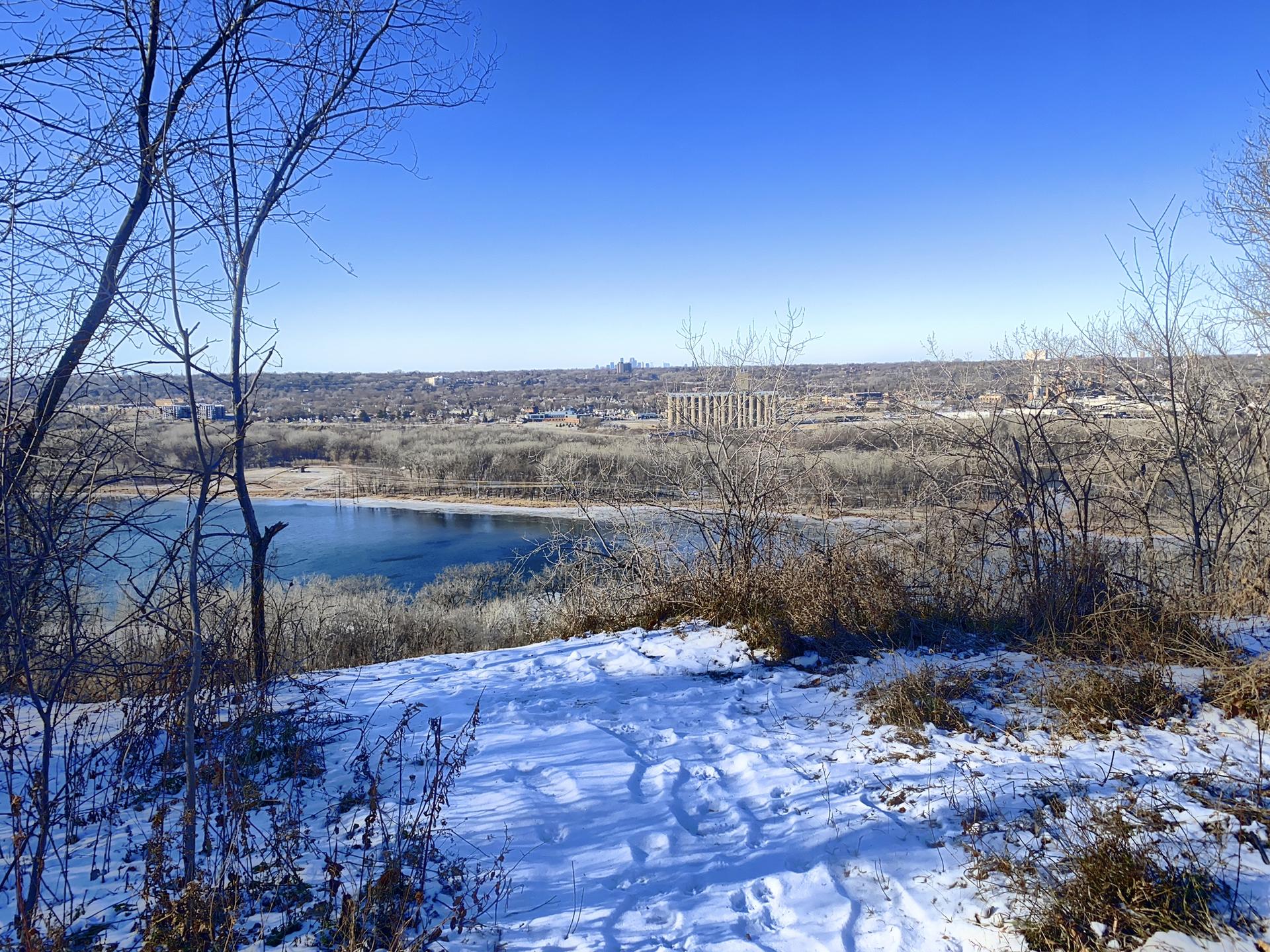 Snowy landscape with bare trees overlooking a river and distant buildings.