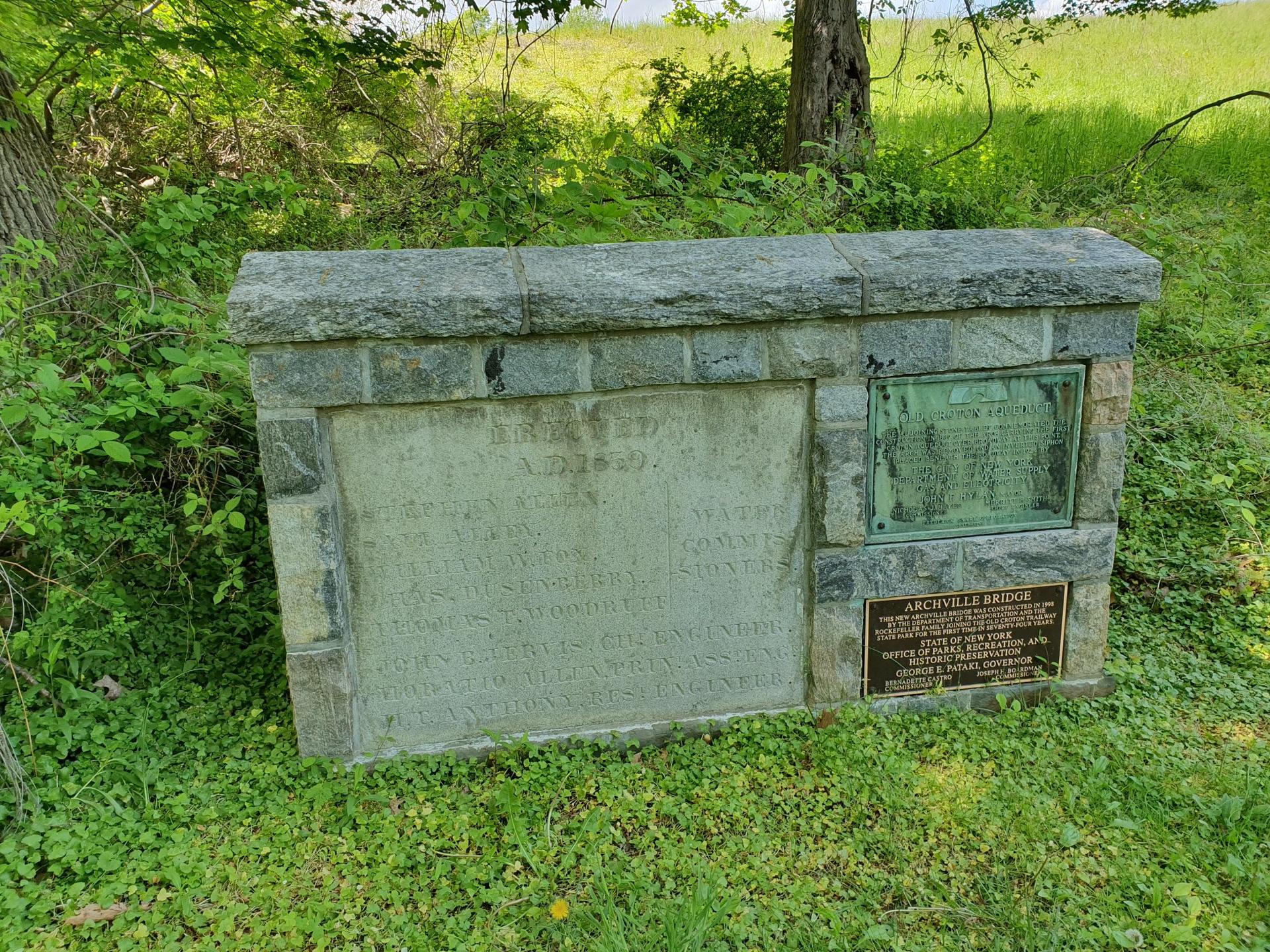 Stone monument with engraved plaques, surrounded by green grass and foliage.